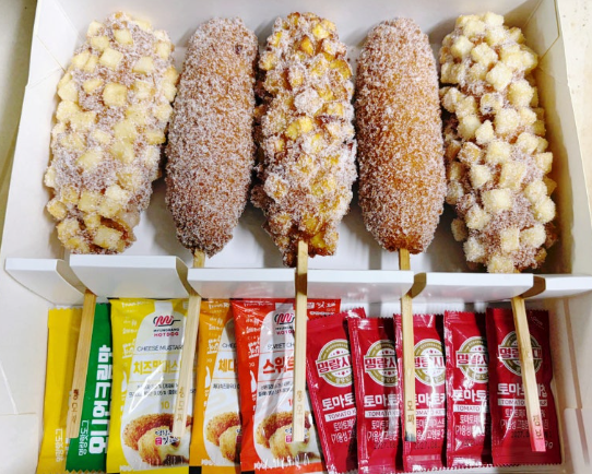 A box of Korean corn dogs from Myeongrang Hot Dog (명랑핫도그), a popular Korean hot dog chain. The box includes different varieties, some coated with diced potatoes and others covered in breadcrumbs. Ketchup and mustard packets are provided for dipping or topping.