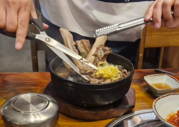 At a Korean restaurant, kitchen scissors (음식용 가위) and tongs (집게) are used to cut the beef ribs in a bowl of galbitang (갈비탕).