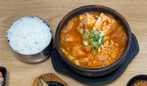 A pot of kimchi jjigae (김치찌개) with tofu and green onions is served alongside a bowl of rice. These days, some Korean restaurants add ramen noodles (라면사리) to kimchi jjigae (김치찌개), serving the hearty stew with a bowl of rice.