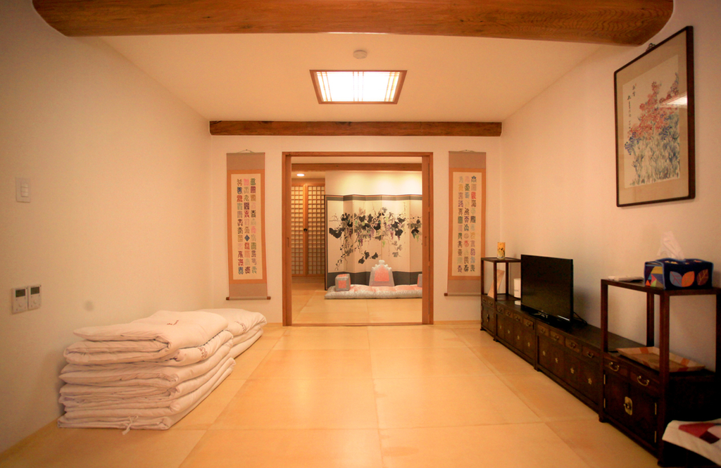 A traditional Korean-style room features a low wooden table, floor cushions, and neatly stacked bedding. Wooden furniture and traditional artwork decorate the room, while sliding doors lead to another room. This space reflects the traditional Korean lifestyle of sitting and sleeping on the floor.